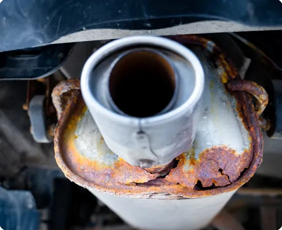 Close-up view of a rusty, corroded car exhaust pipe and muffler, showing significant damage and deterioration on the metal surface. Exhaust Repair In Millersport, OH At Kreager Tire and Service