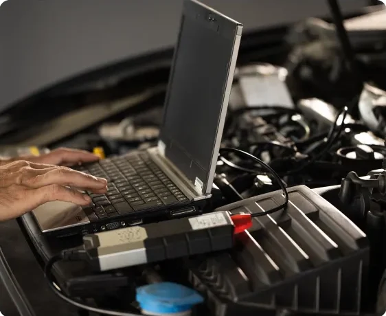 A person uses a laptop connected to a car’s engine for diagnostics or repairs, with hands on the keyboard and the vehicle hood open, showing engine components. Engine Diagnostics & Repair In Millersport, OH At Kreager Tire and Service