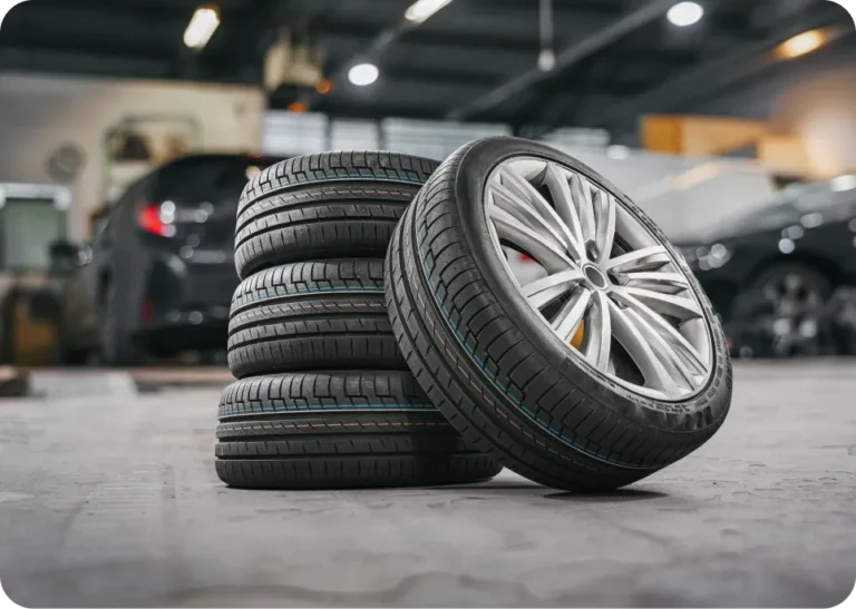 Tire Services In Millersport, OH At Kreager Tire and Service. Four new car tires, one mounted on a shiny alloy wheel, are stacked together on the floor of an auto repair shop. Other vehicles and equipment are visible blurred in the background.