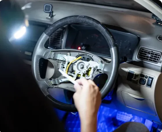 A person holds wires inside a car with the steering wheel cover removed, exposing internal components. The car dashboard and pedals are visible in the background, illuminated by a bright light. Steering and Suspension In Millersport, OH At Kreager Tire and Service