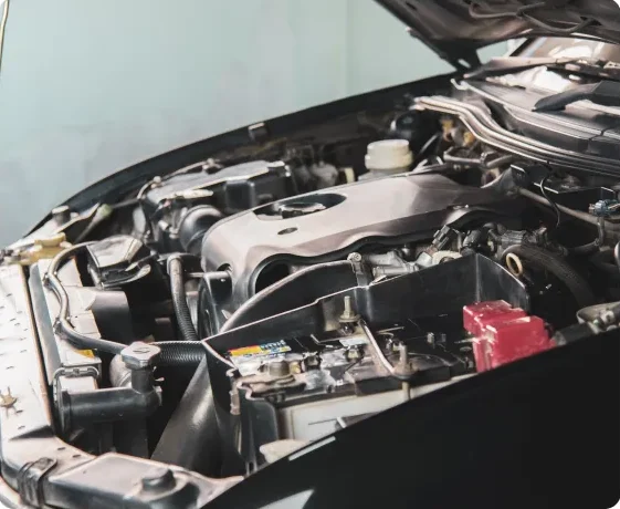 A close-up view of a car engine with the hood open, showing various engine components, including the battery, hoses, and engine cover, in a well-lit workspace. Vehicle Maintenance In Millersport, OH At Kreager Tire and Service
