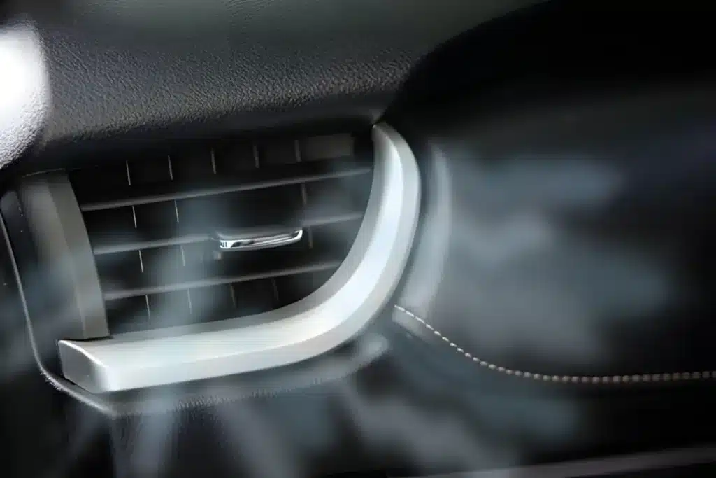 Close-up of a car air vent with cool air visibly blowing out. The vent is surrounded by a silver trim and set against a dark dashboard with visible stitching.