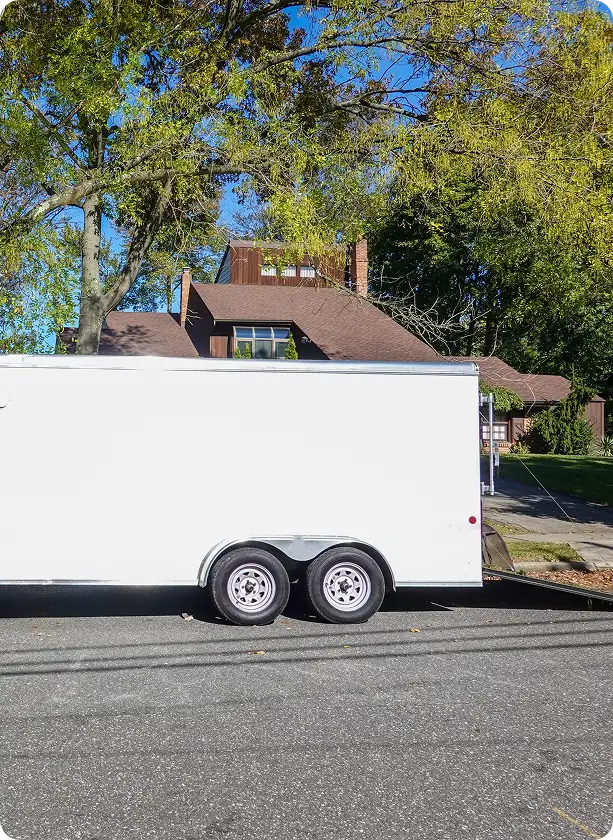 A white enclosed trailer is parked on a street in front of a house partially obscured by trees with green leaves, under a clear blue sky. Trailer Tire In Millersport, OH At Kreager Tire and Service