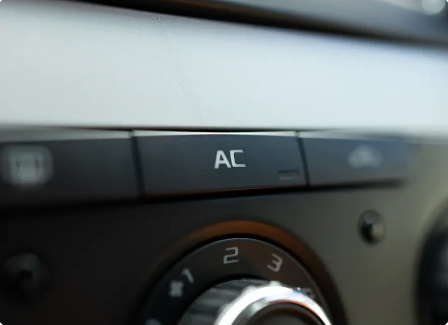 Close-up of a car dashboard showing an AC button for air conditioning above a circular dial with numbers. The image is slightly blurred and focused on the AC button. AC & Heater Repair In Millersport, OH At Kreager Tire and Service