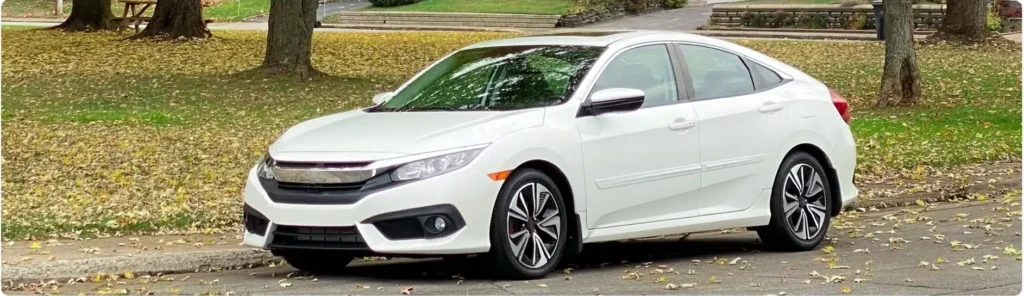 A white sedan car is parked on the side of a leaf-covered street in a park-like setting, with trees and grass in the background. Honda Repair In Millersport, OH At Kreager Tire and Service