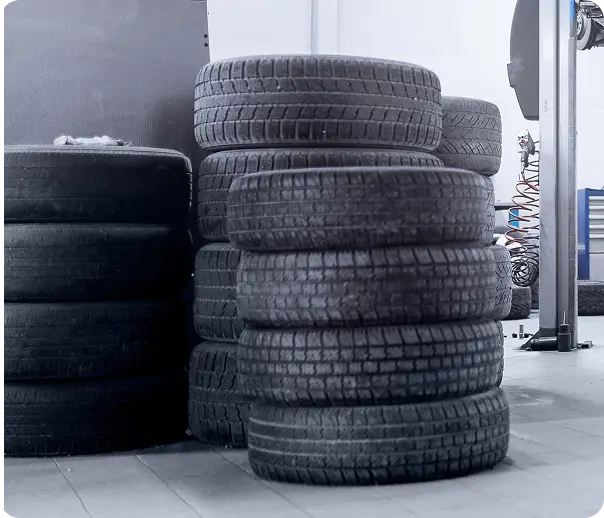 Stacks of black car tires are neatly arranged on a tiled floor in an indoor garage or workshop, with some automotive equipment visible in the background. Tire Services In Millersport, OH At Kreager Tire and Service