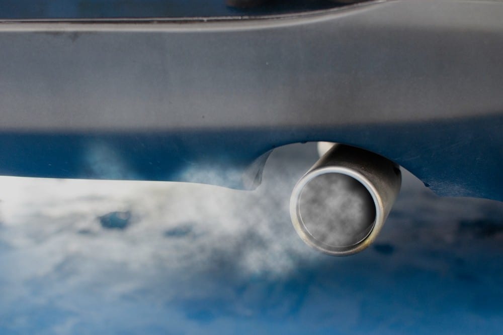 Metallic vehicle exhaust pipe emitting visible smoke against a blurred blue background, suggesting engine activity or combustion issues.